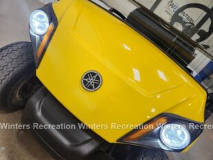 Close-up of a yellow vehicle front with headlights and a logo, showcasing a sleek design and rugged tires.